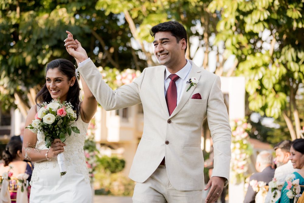 A celebratory couple in matching white attire walk hand in hand down the aisle at an outdoor wedding ceremony.