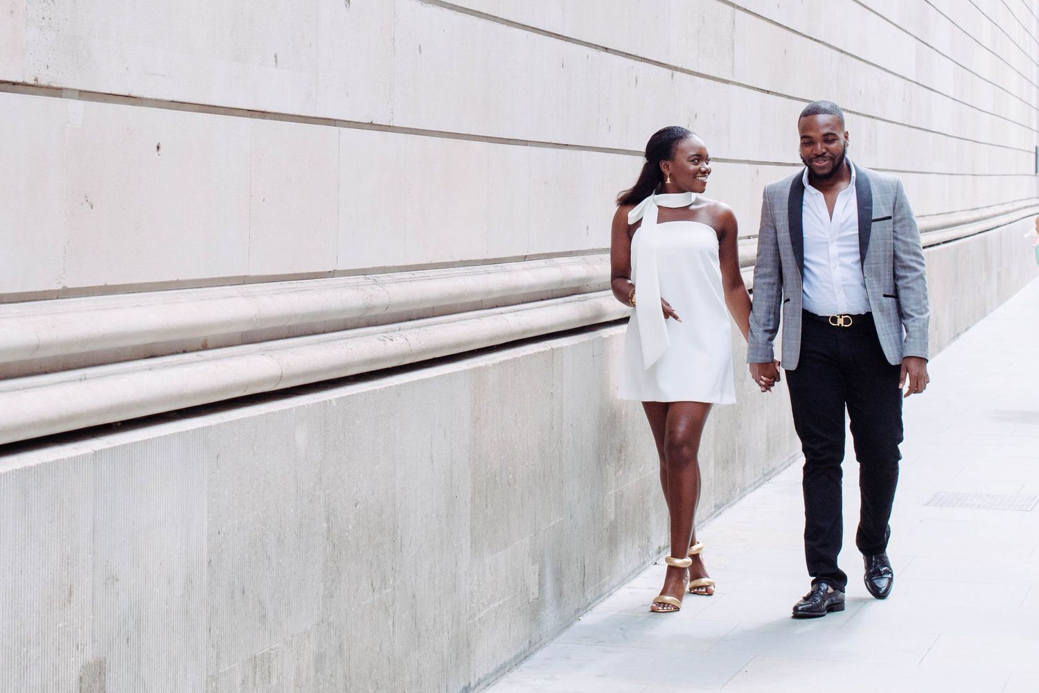 Engagement session in London, the couple is walking down a street on a sunny day. She is wearing a beautiful white dress,