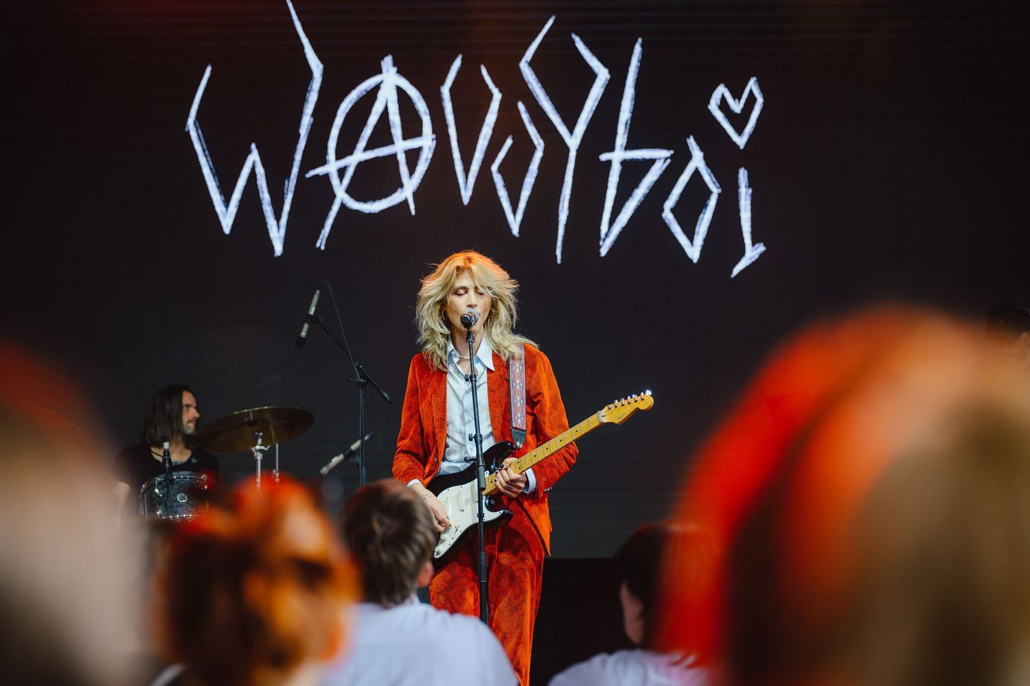 Performer playing electric guitar on stage with glowing text display and red lighting.