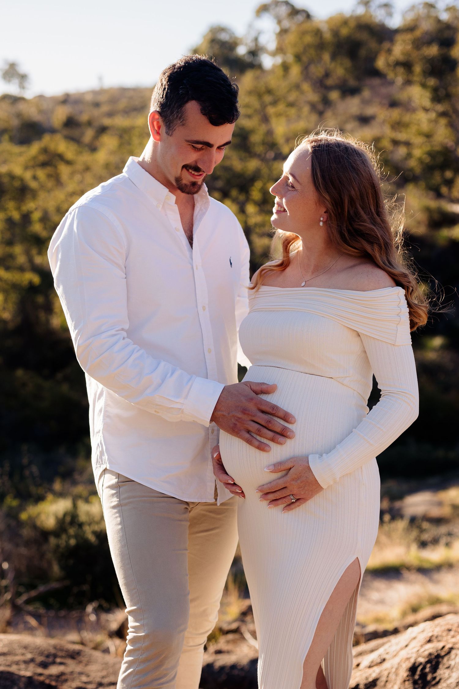 A couple tenderly cradles a pregnant belly outdoors, wearing white and cream outfits against a sunlit natural backdrop.