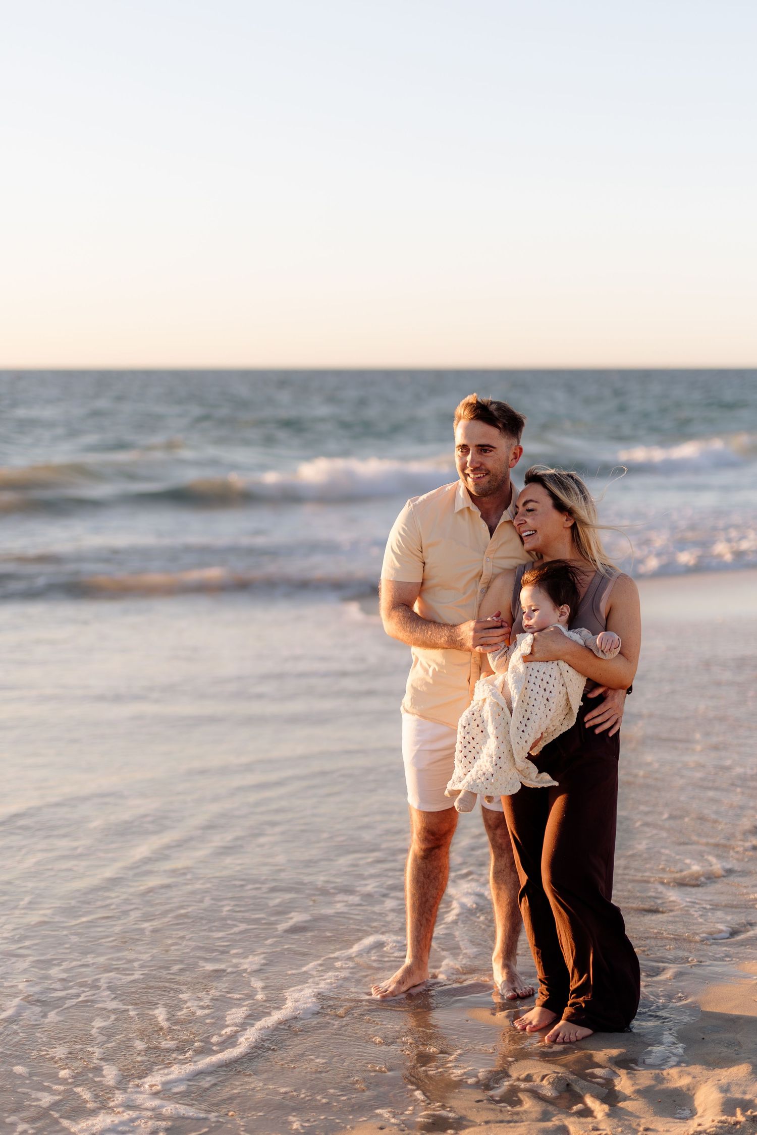 Happy family of three enjoying a sunset beach walk, parents smiling while holding their young child by the ocean waves.