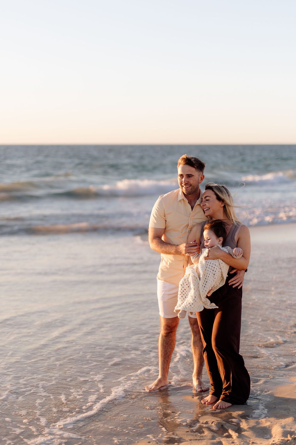 Happy family of three enjoying a sunset beach walk, parents smiling while holding their young child by the ocean waves.