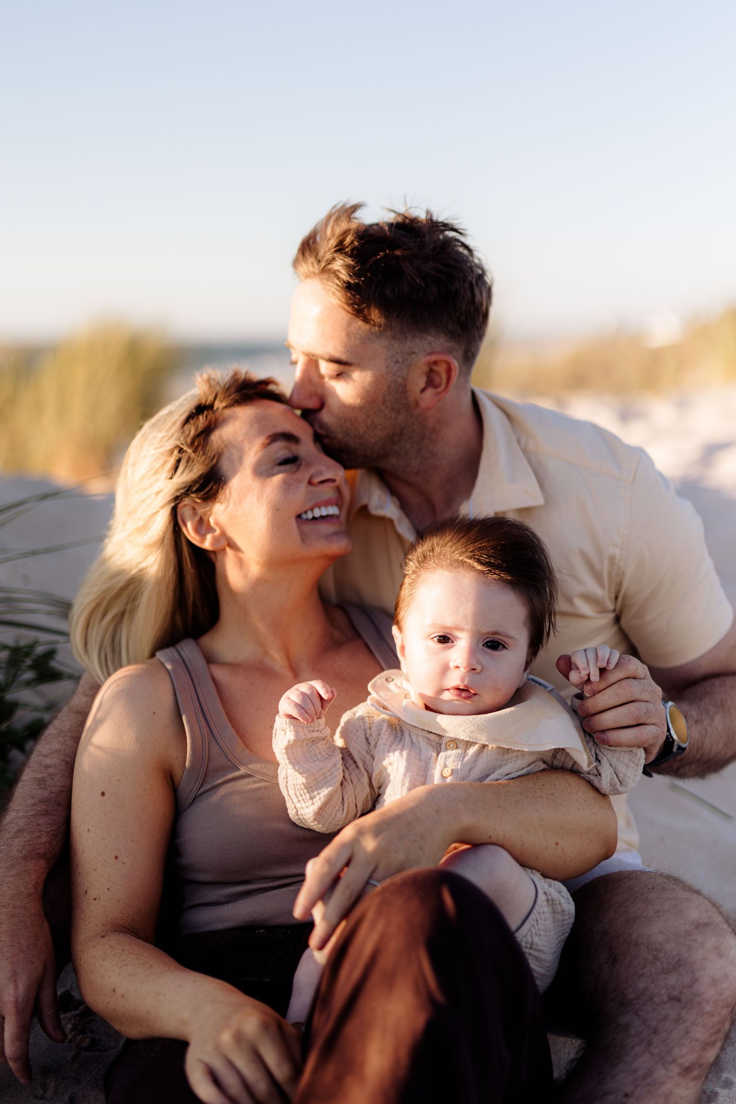 A happy family of three sits on a sandy beach, with a man kissing a laughing woman while she holds their baby.