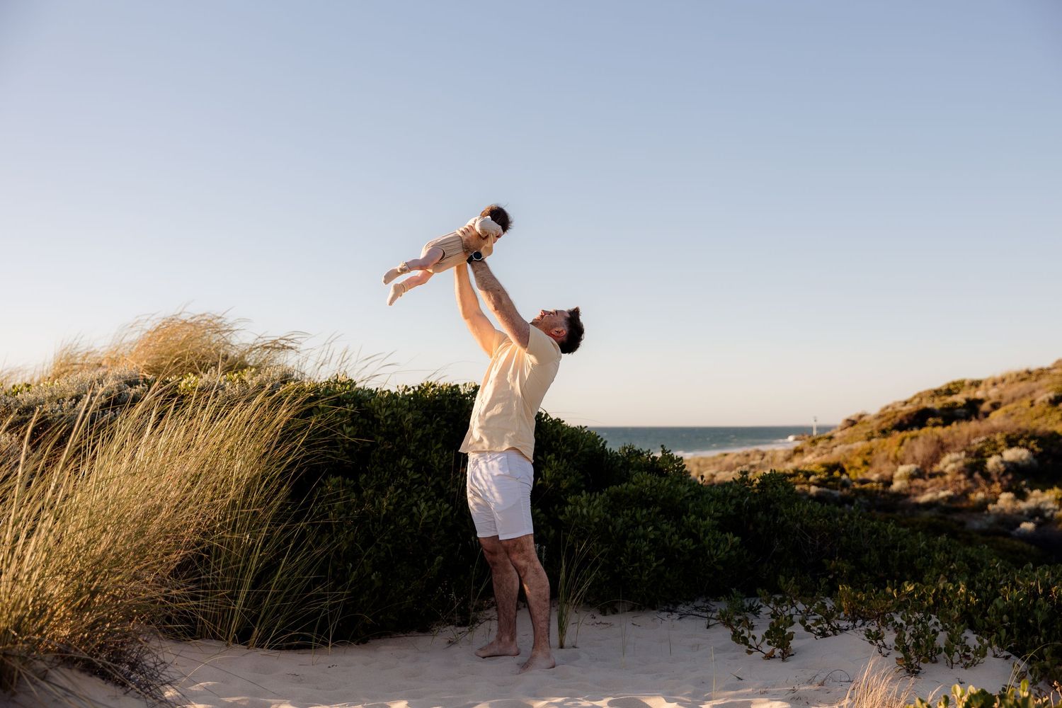Father joyfully lifting baby in the air on a sunny beach with green dunes and ocean in the background.