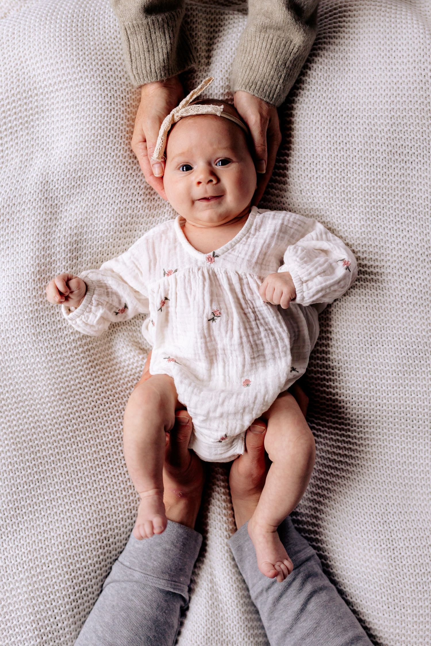 Adorable baby girl in white floral onesie and headband lying on blanket, held gently by parent hands.