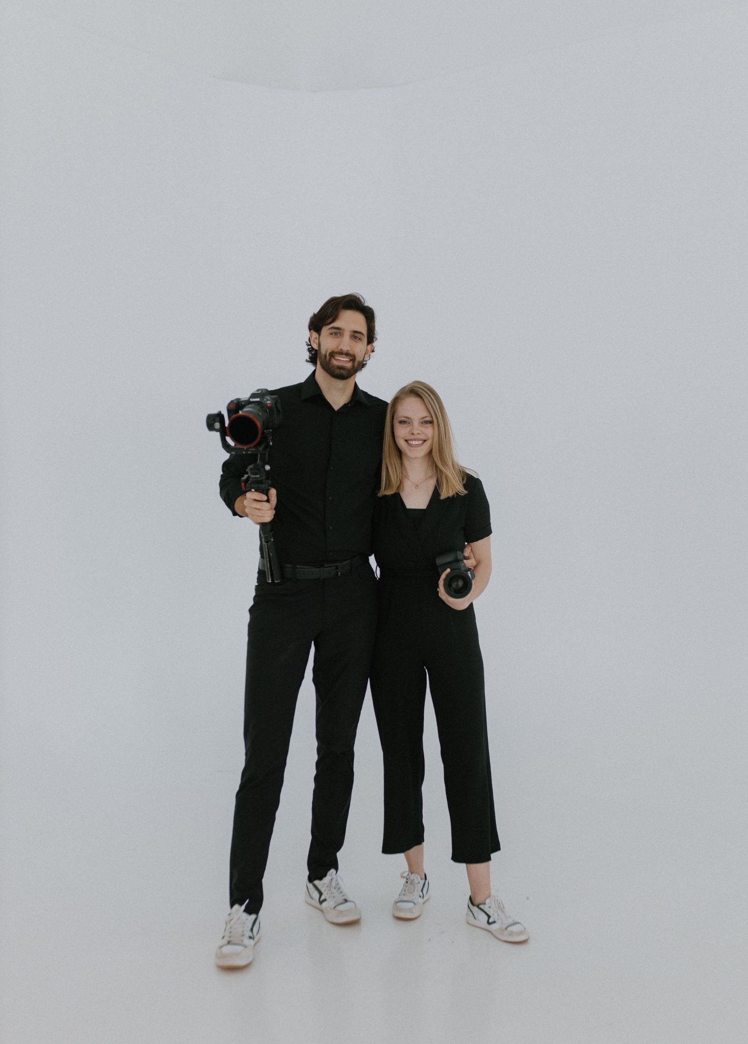 Two photographers in matching black outfits pose together against a white backdrop with professional cameras.