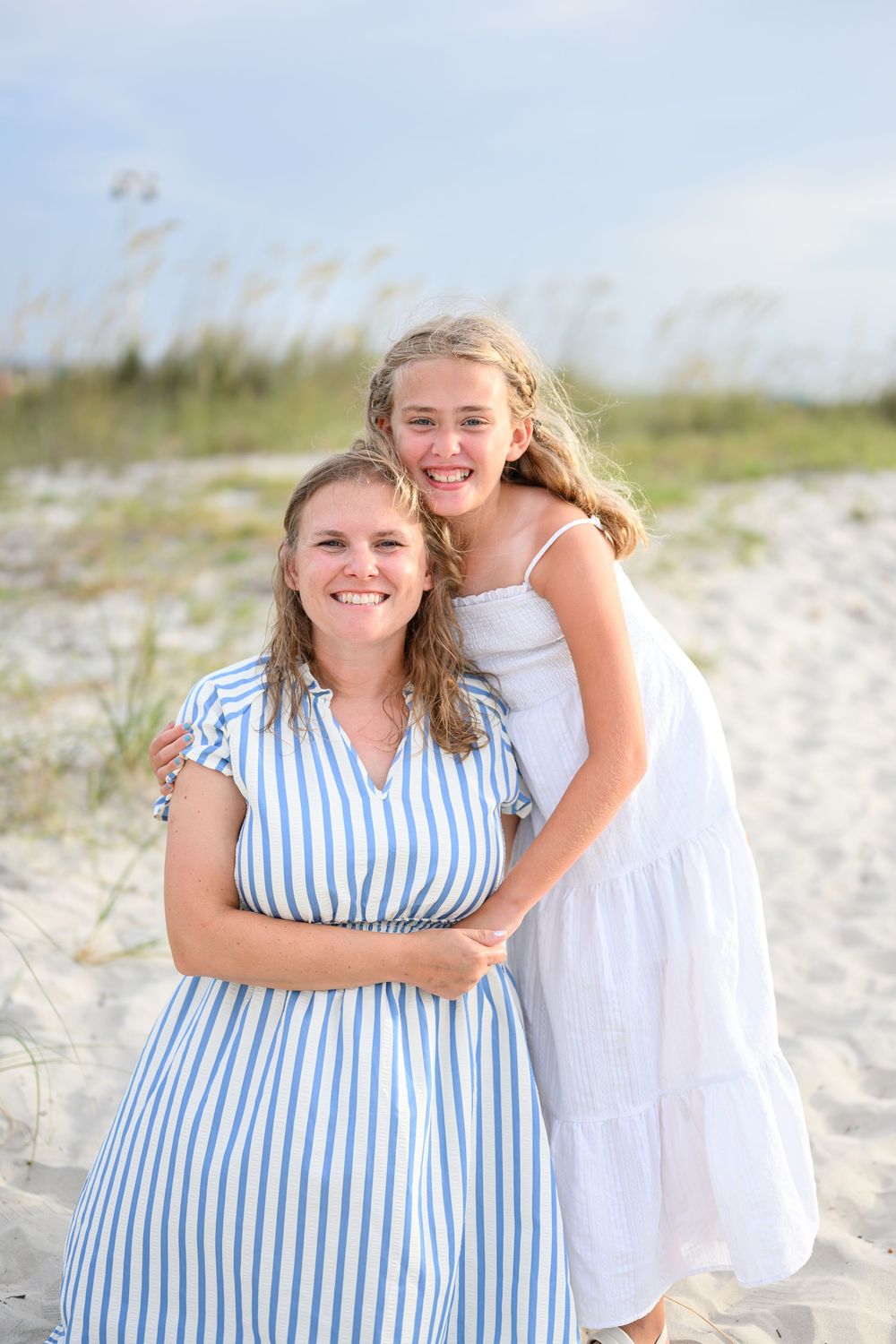 A woman and young girl in matching blue and white outfits smile together on a sandy beach.
