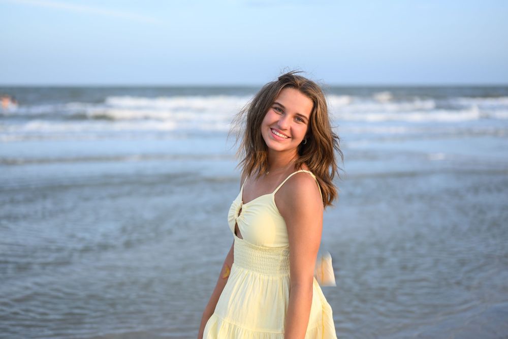 Young woman in a yellow sundress smiling at the beach with ocean waves in the background.