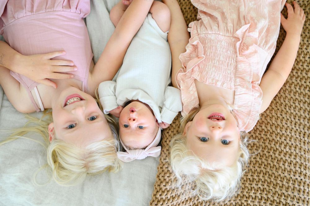 Three children in light pastel outfits lie on a carpet together, looking upward with happy expressions.