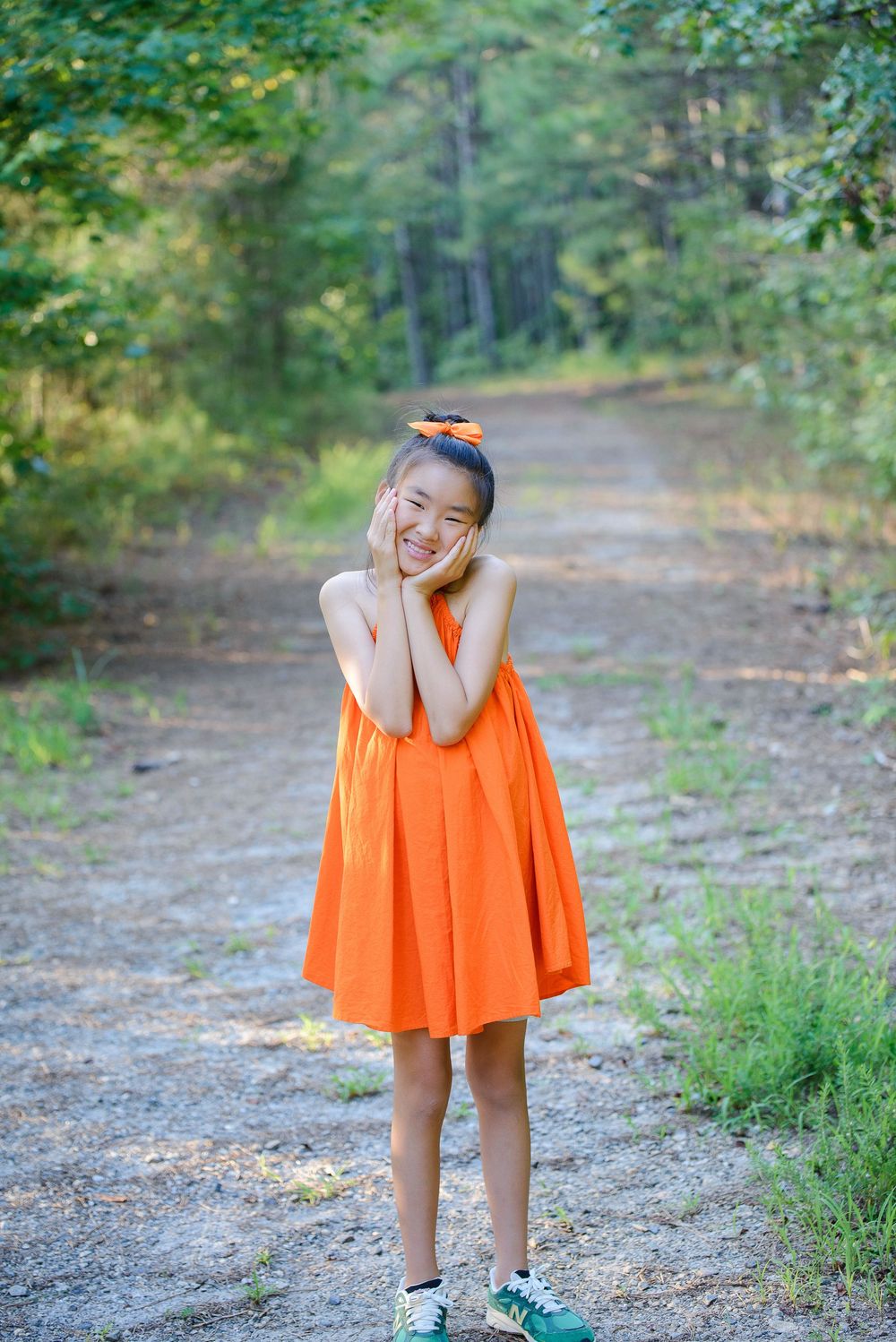 A person in a bright orange dress and headband stands playfully on a forest path on a sunny summer day.
