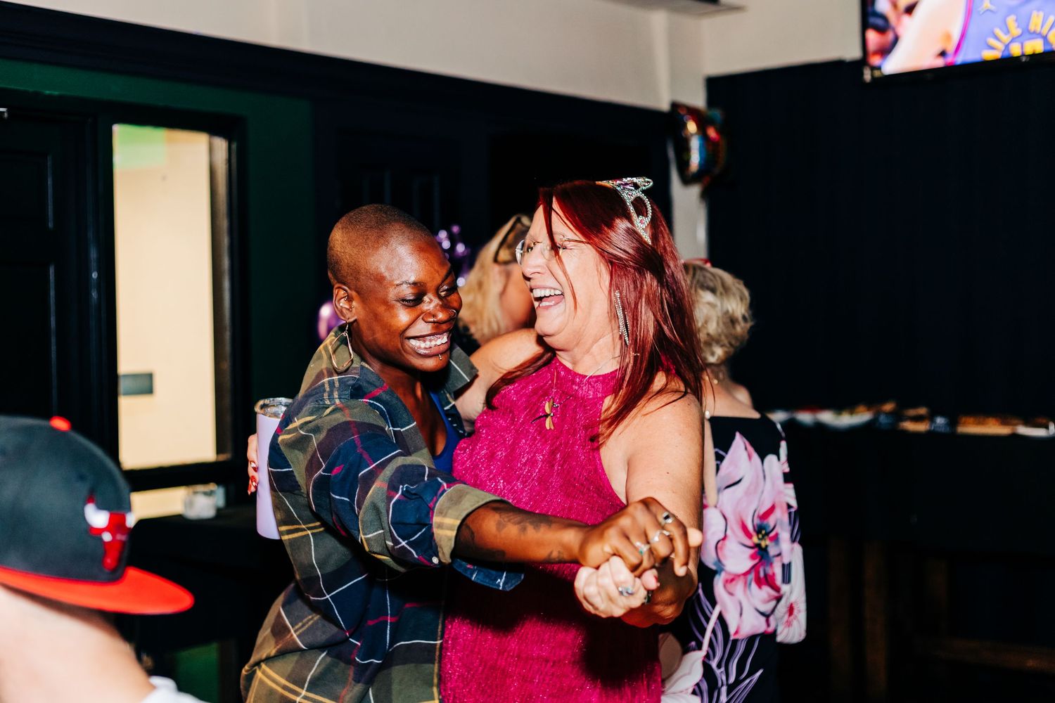 Two people dancing together joyfully at a party, with one wearing a denim jacket and another in a bright pink dress at an event.