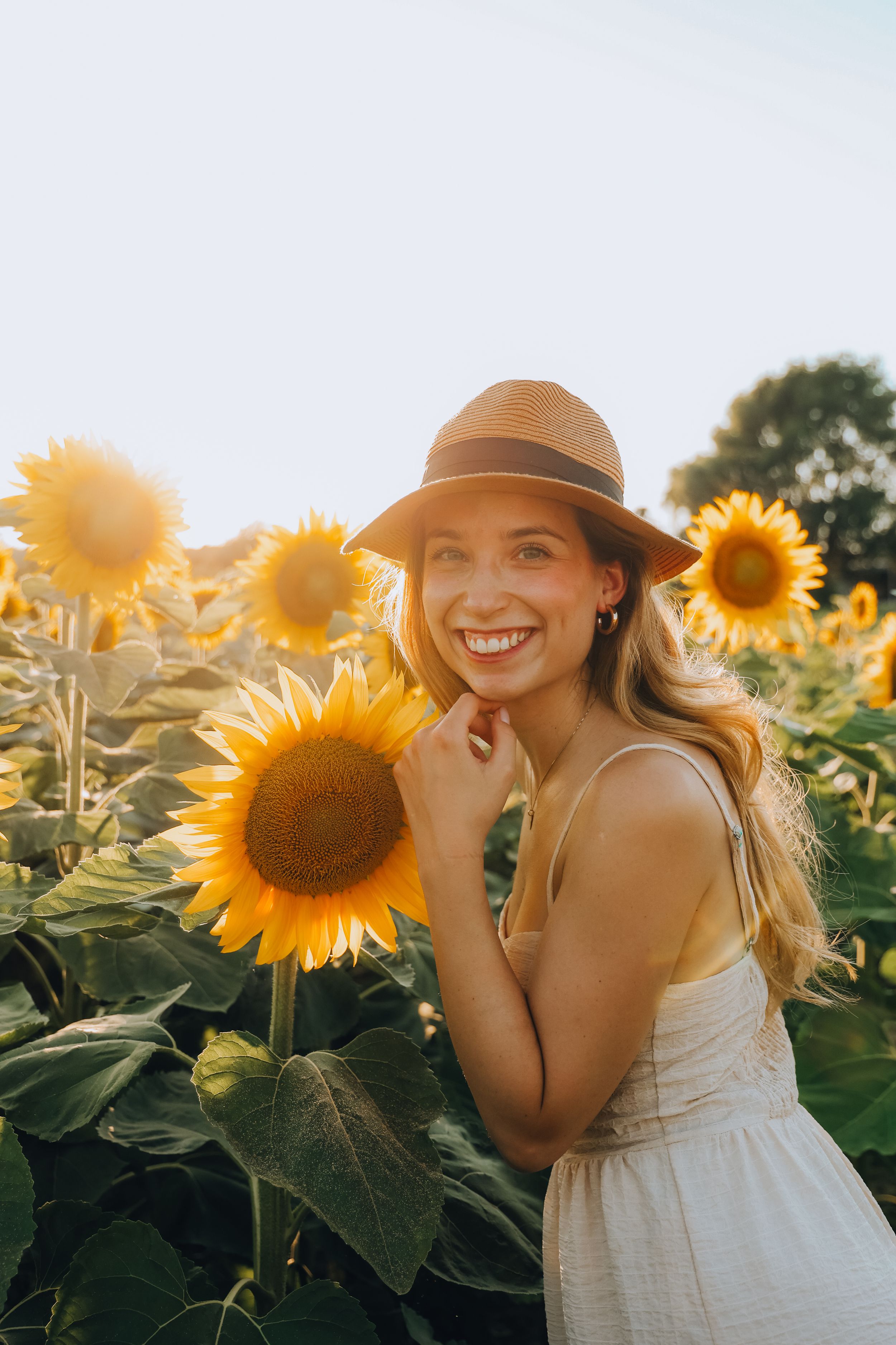 Smiling blonde woman in hat posing beside bright sunflower in sunny field.