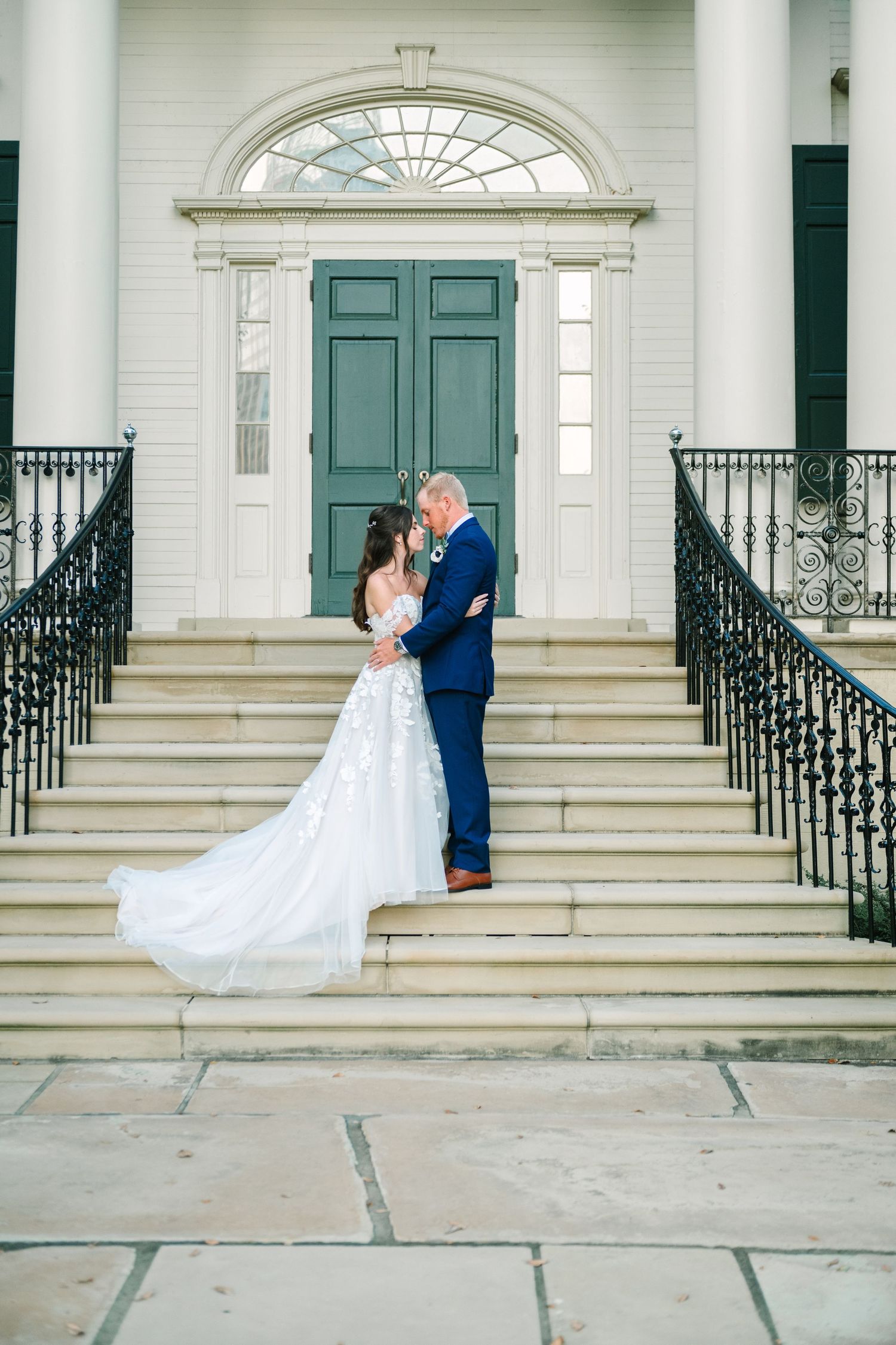 A couple shares a tender moment in front of a rustic white barn with a mobile bar cart nearby at their wedding celebration.