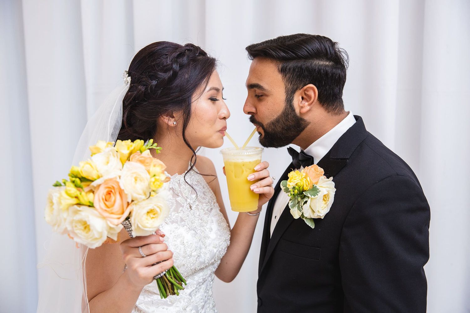 A bride and groom drinking from the same glass of drinking juice with straws, standing against a white curtain