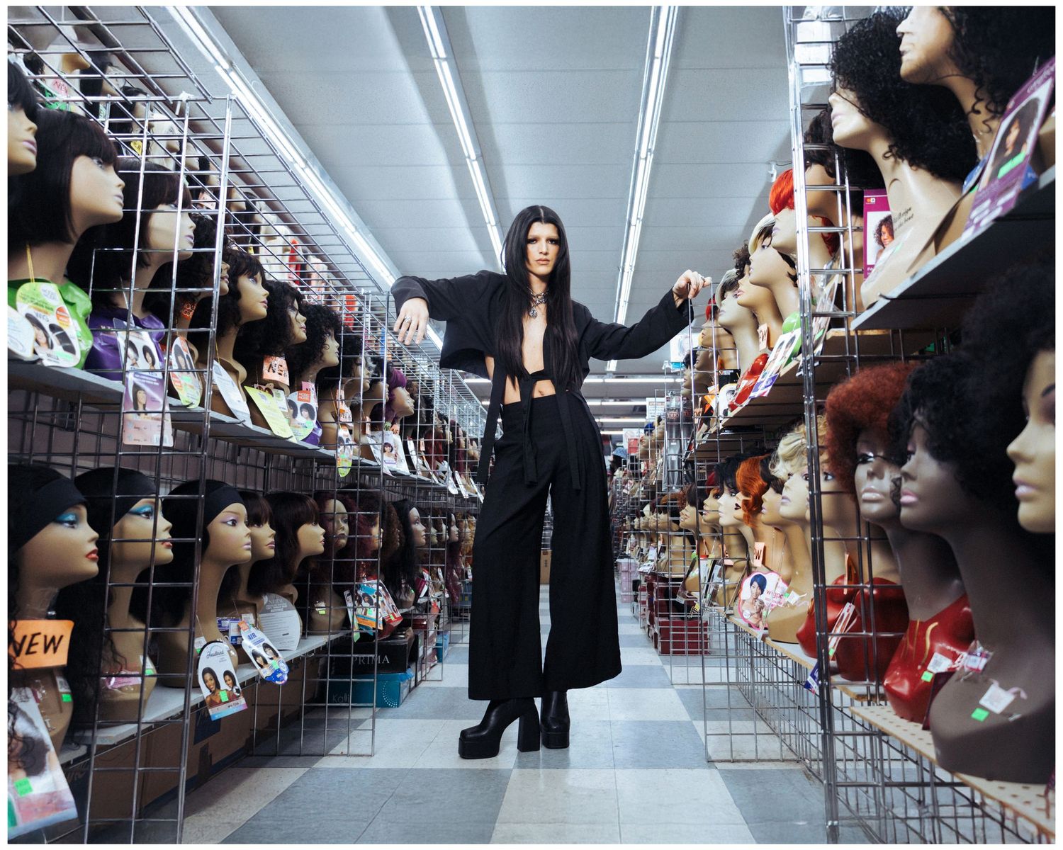A figure in black clothing stands dramatically between aisles filled with various wigs on display shelves in a retail store.