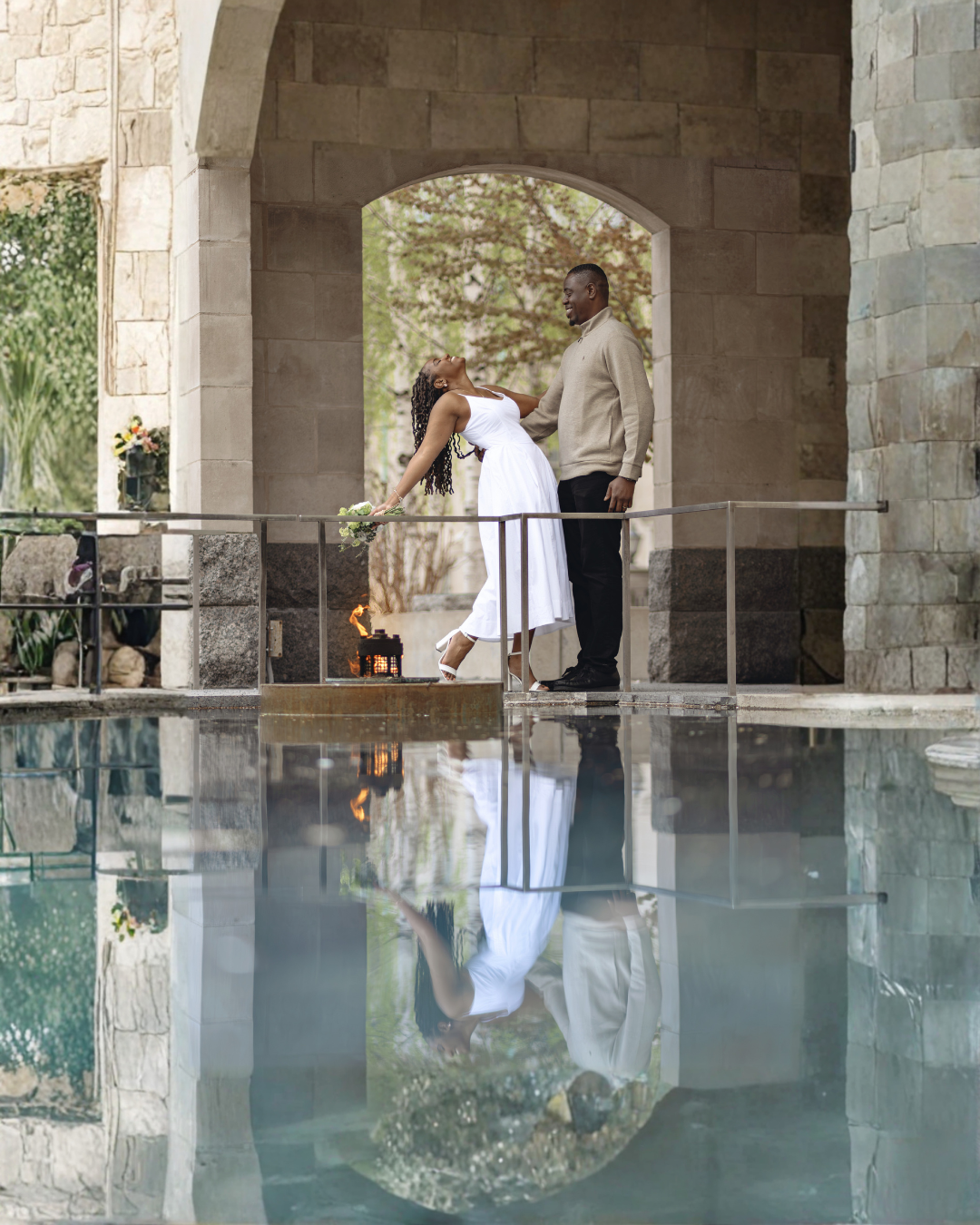 A couple shares a romantic moment under a stone arch reflected in a still pool of water.