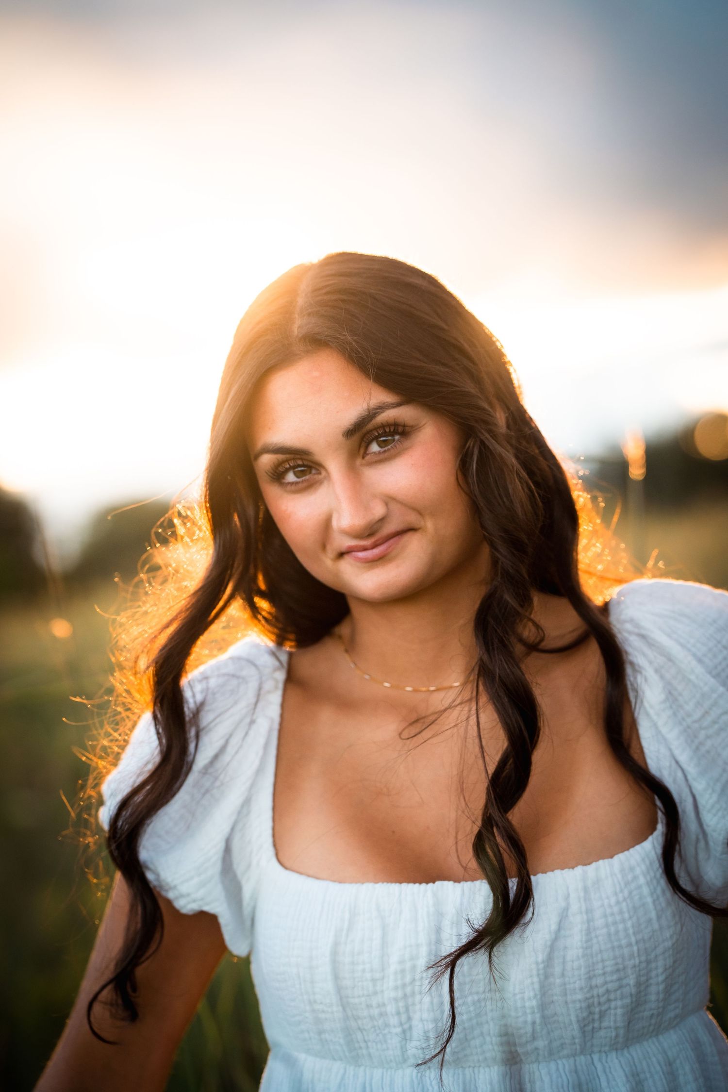 A woman in a light blue dress poses outdoors during golden hour with sunlit hair and warm lighting.