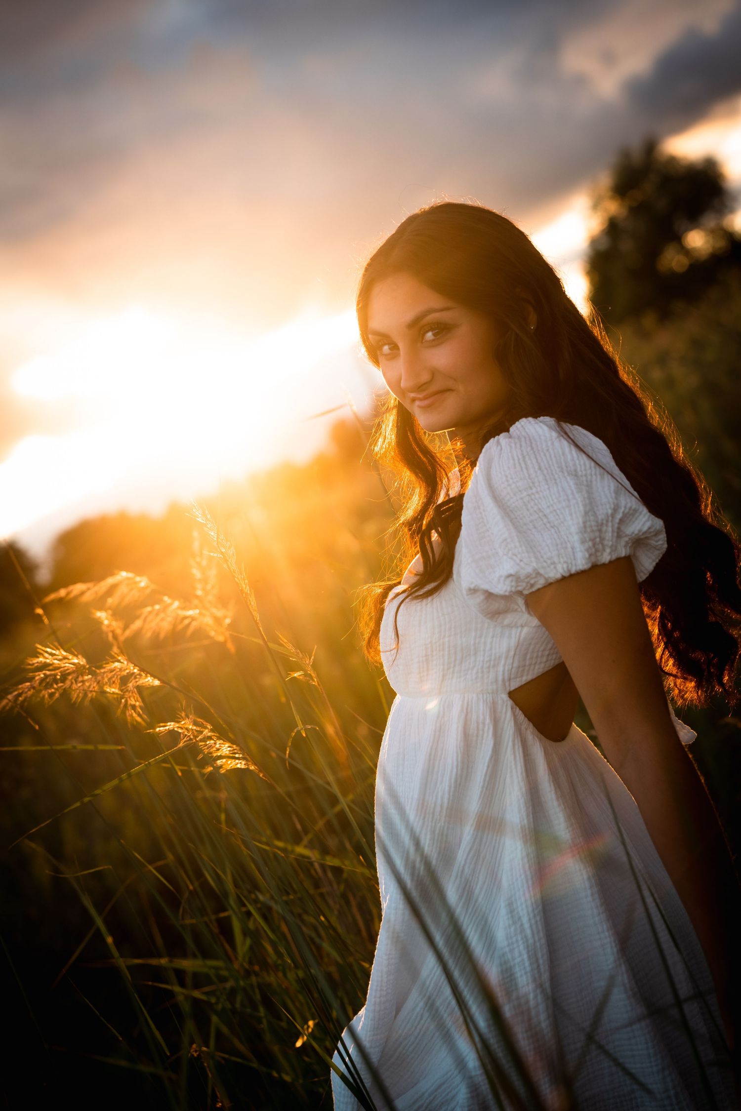 A woman in a white dress poses in a field at golden hour as sunlight streams through tall grass and creates a warm glow.