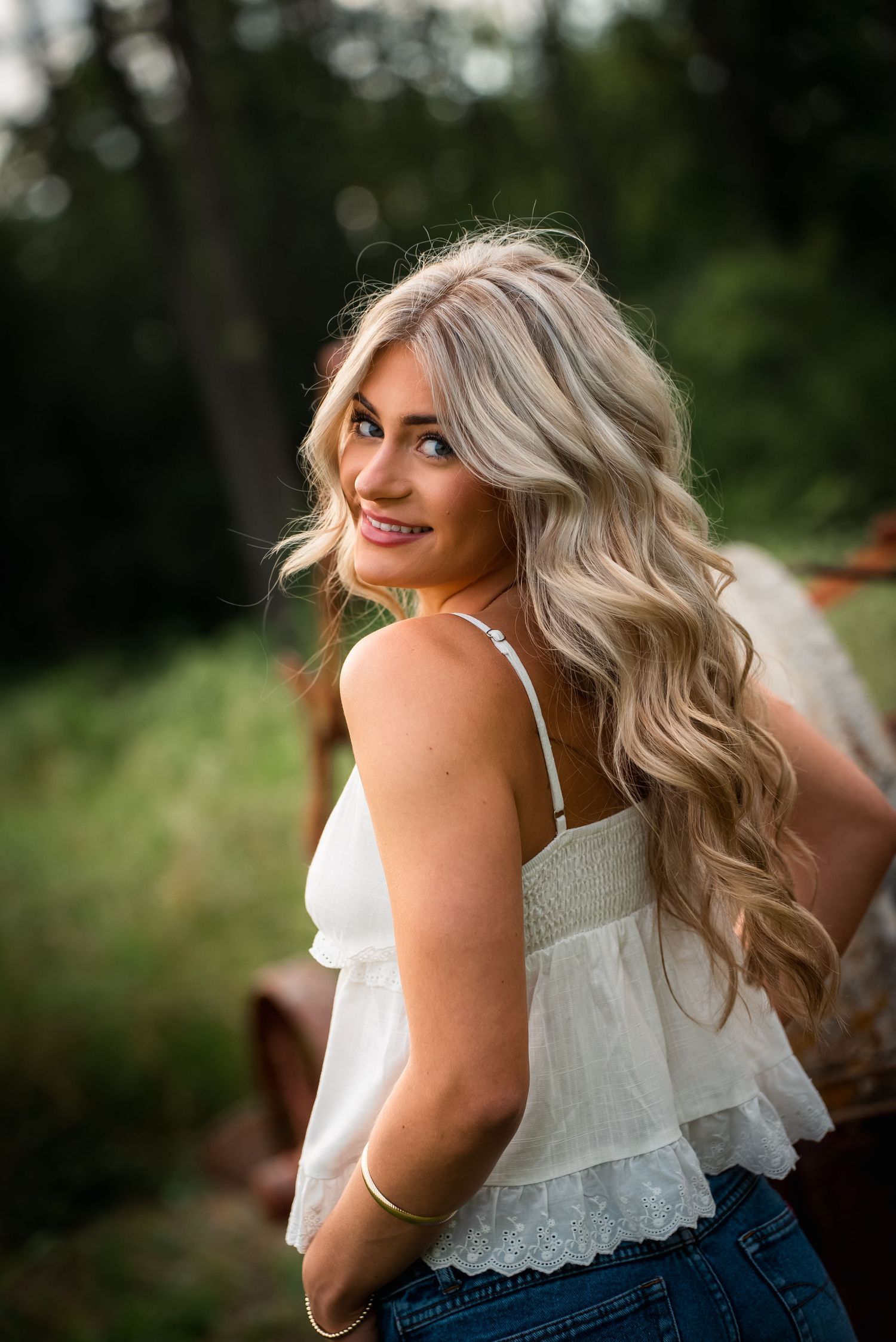 A person with wavy blonde hair and a white lace top poses outdoors against a blurred natural background.
