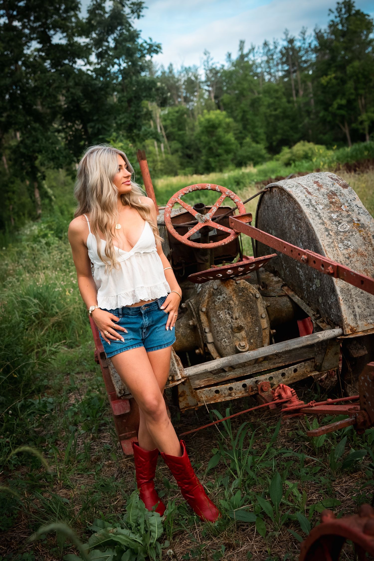 A rustic country photoshoot featuring a white top, denim shorts and red boots next to vintage farm equipment.