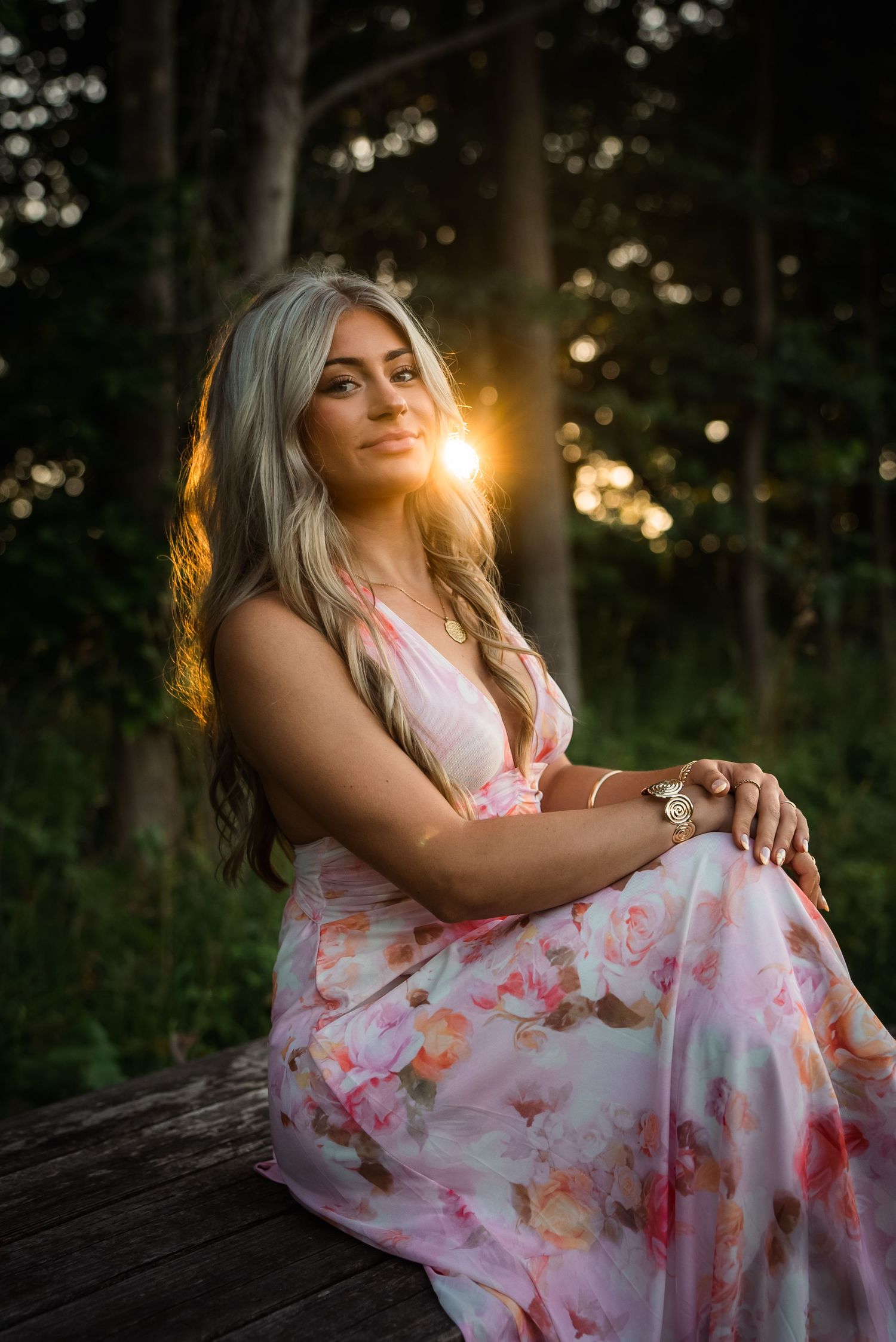 A person in a floral dress sits on a wooden surface at sunset with golden light filtering through trees in the background.