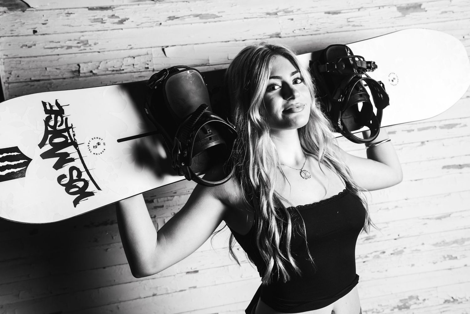 Black and white photo of someone posing with a snowboard and headphones against a wooden wall.