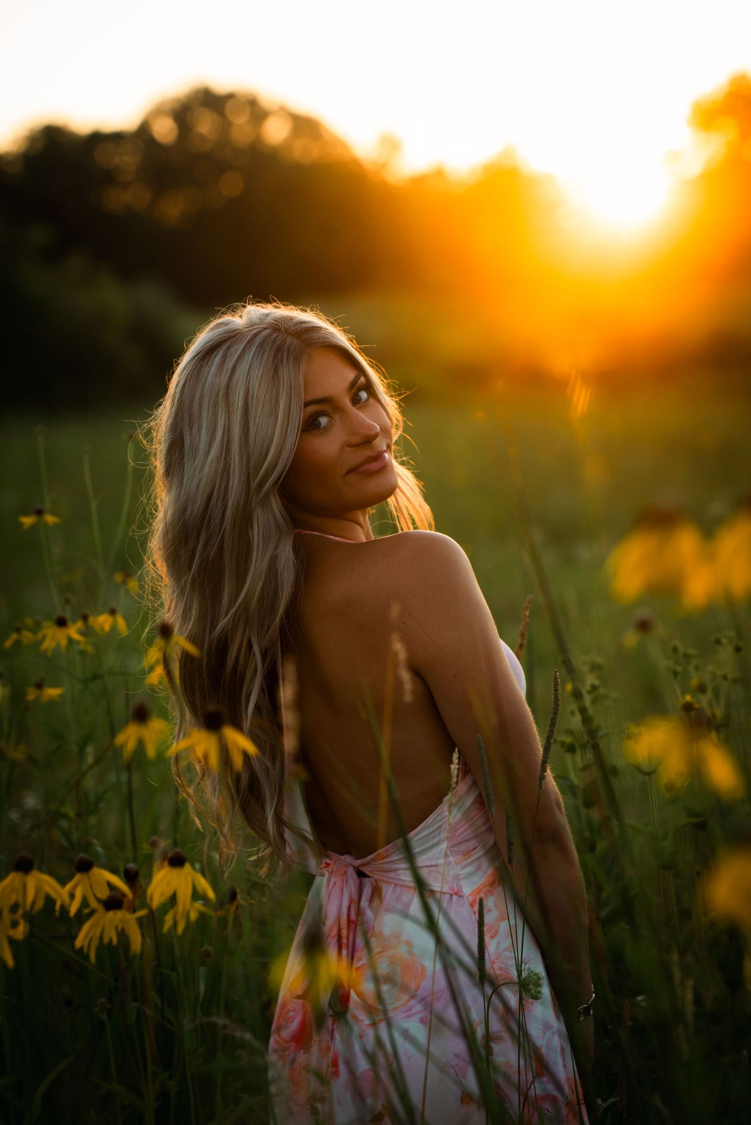 A backlit figure in a floral dress poses in a wildflower meadow at golden hour sunset.
