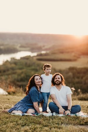 Family sits together on blanket overlooking scenic river valley during golden sunset.