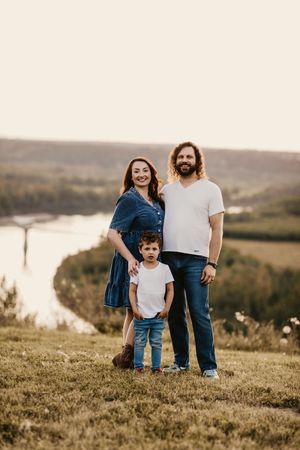Family stands together on grassy hilltop overlooking water during golden hour in nature setting.
