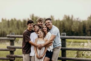 A family laughs and embraces while walking on a wooden boardwalk surrounded by tall grass and trees.