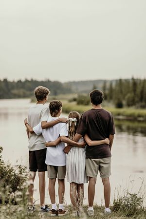 Four people stand together with arms around each other looking out at a serene lake surrounded by trees at dusk.
