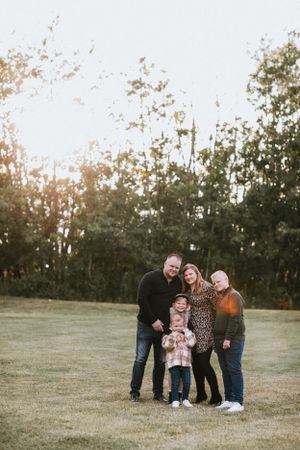 A family poses together on a grassy field at sunset with trees in the background.