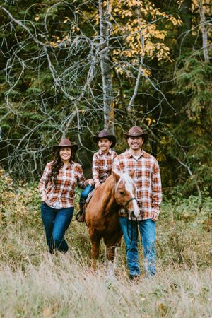 Three people wearing plaid shirts and cowboy hats stand outdoors with a brown horse in a grassy field near autumn trees.