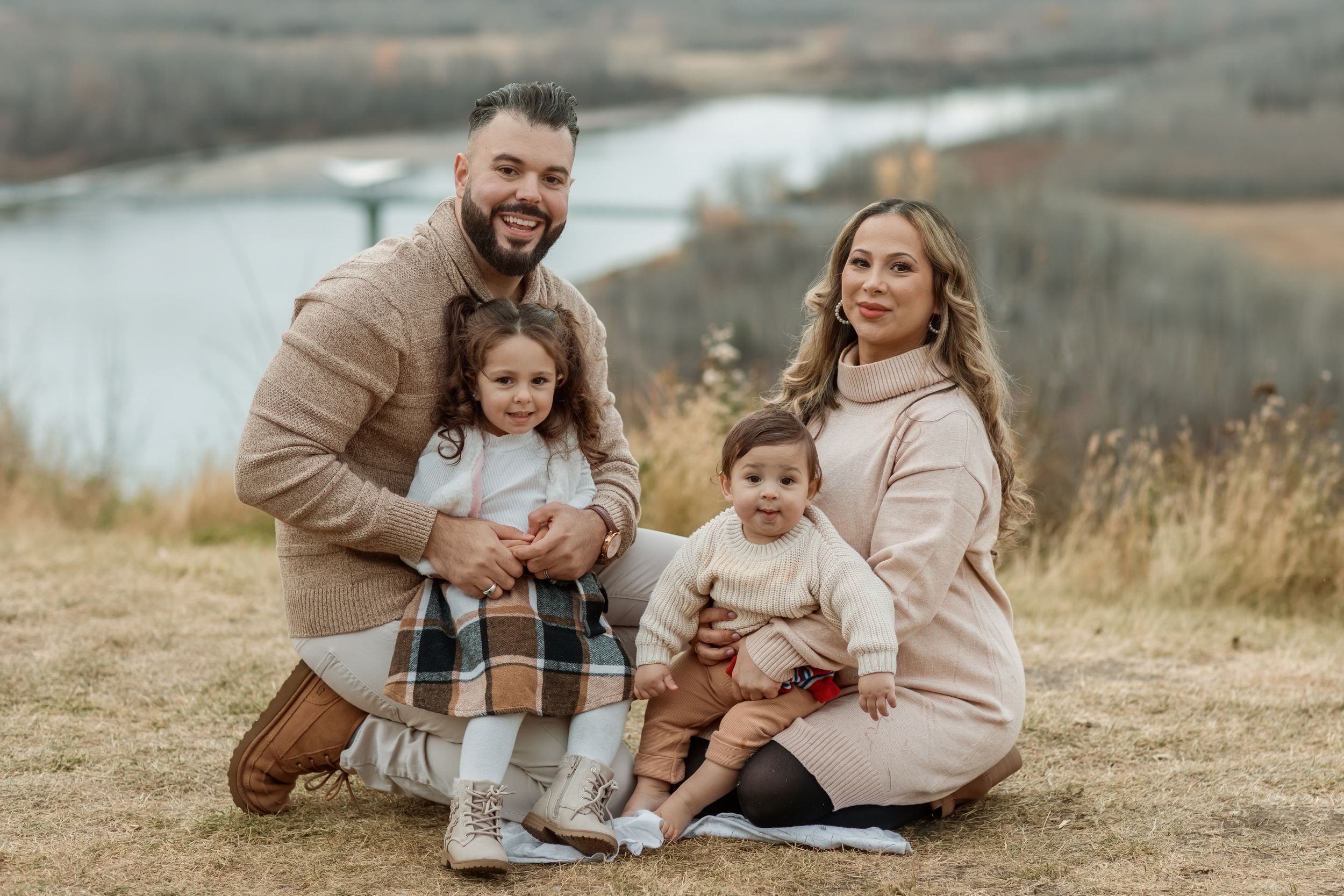 A family in warm earth tones sits on a grassy hillside during golden hour with scenic mountain views in the background.