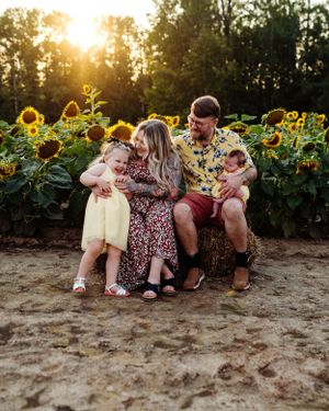 A family sits together in a sunflower field during golden hour sunset, creating a warm and joyful outdoor portrait.