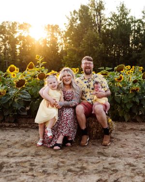 A joyful group sits together in a sunflower field at sunset with golden light filtering through the trees behind them.
