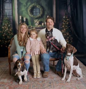 Holiday portrait of a family and their three dogs posing in front of decorated Christmas trees and a wreath.