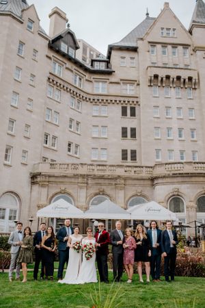A wedding party poses on the lawn in front of a grand historic hotel with arched windows and stone architecture.