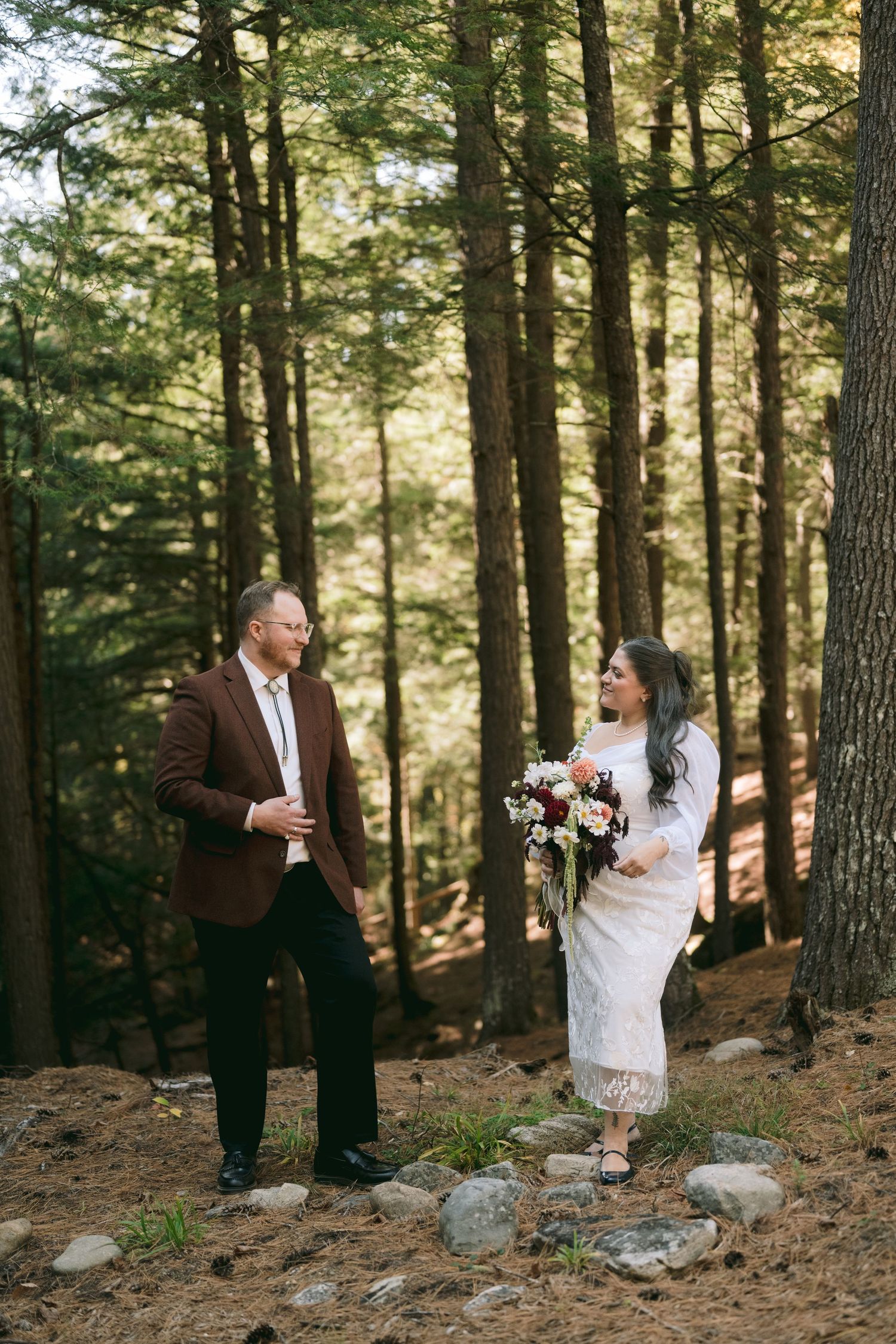 Groom in brown jacket smiles at bride holding a colorful bouquet during a forest elopement ceremony.