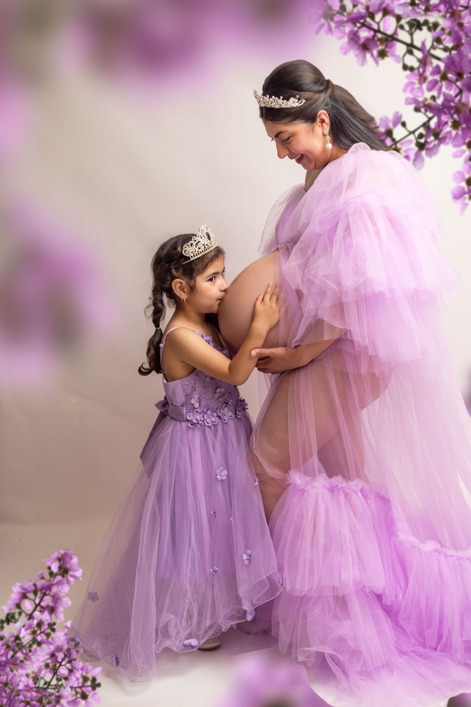 Pregnant woman and young daughter in matching purple tulle gowns and tiaras pose with purple flowers.