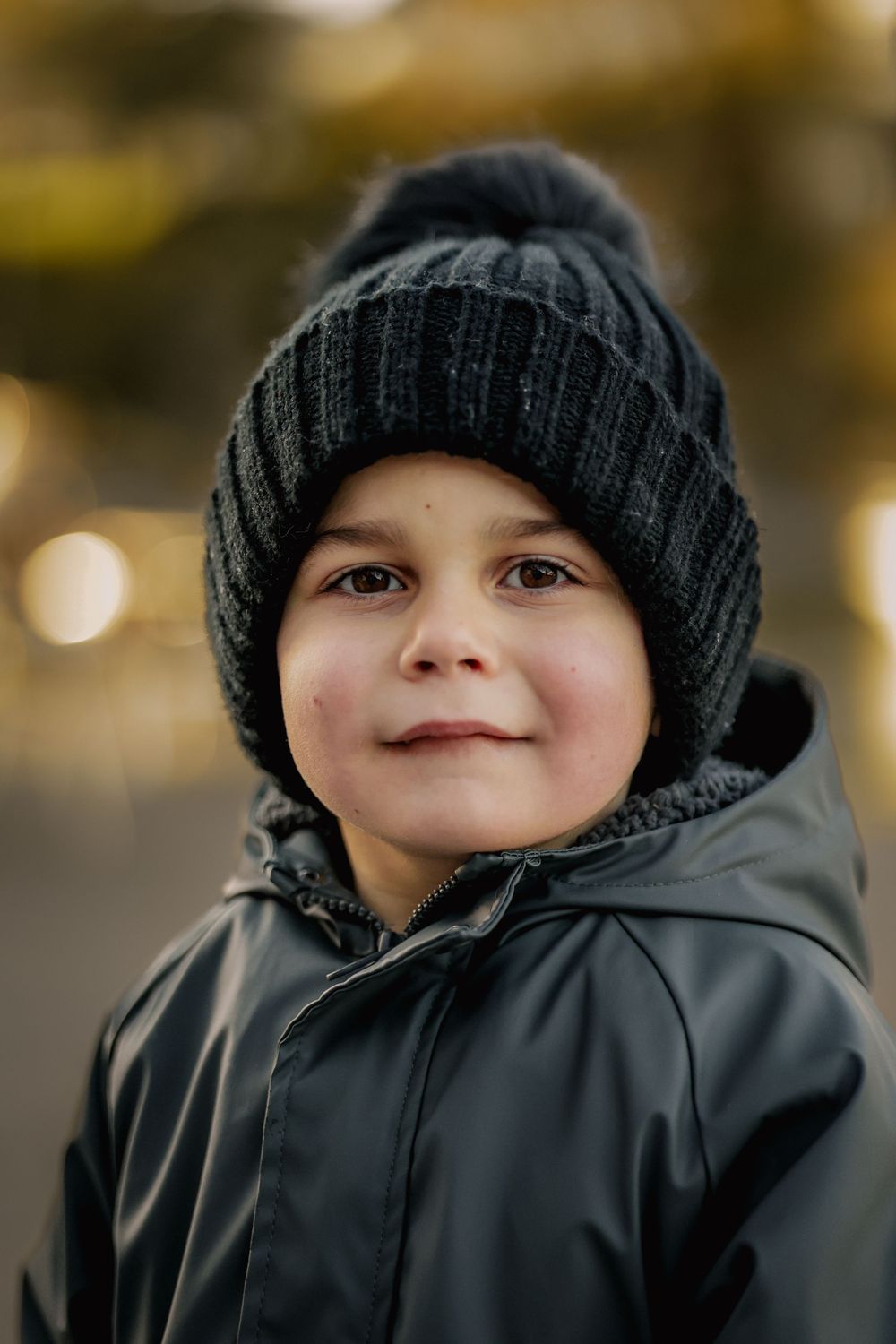 Young child in a dark knit pom-pom beanie and black waterproof jacket smiling softly outdoors against a warm blurred background.