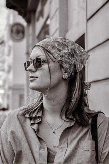 Black and white profile portrait of a woman in sunglasses, a patterned bandana and a button-up shirt, standing on a city street.