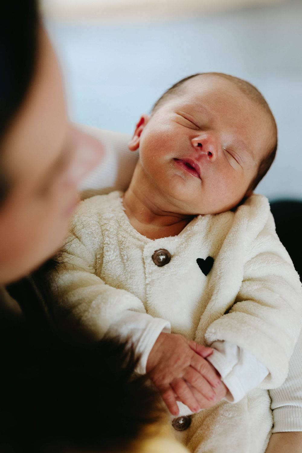 Sleeping newborn baby in a cozy white fleece jacket with a heart patch, resting peacefully in a parent's arms.