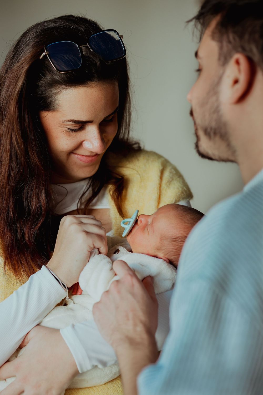 Smiling woman with sunglasses on her head cradles a sleeping newborn baby while a bearded man gently touches the infant.