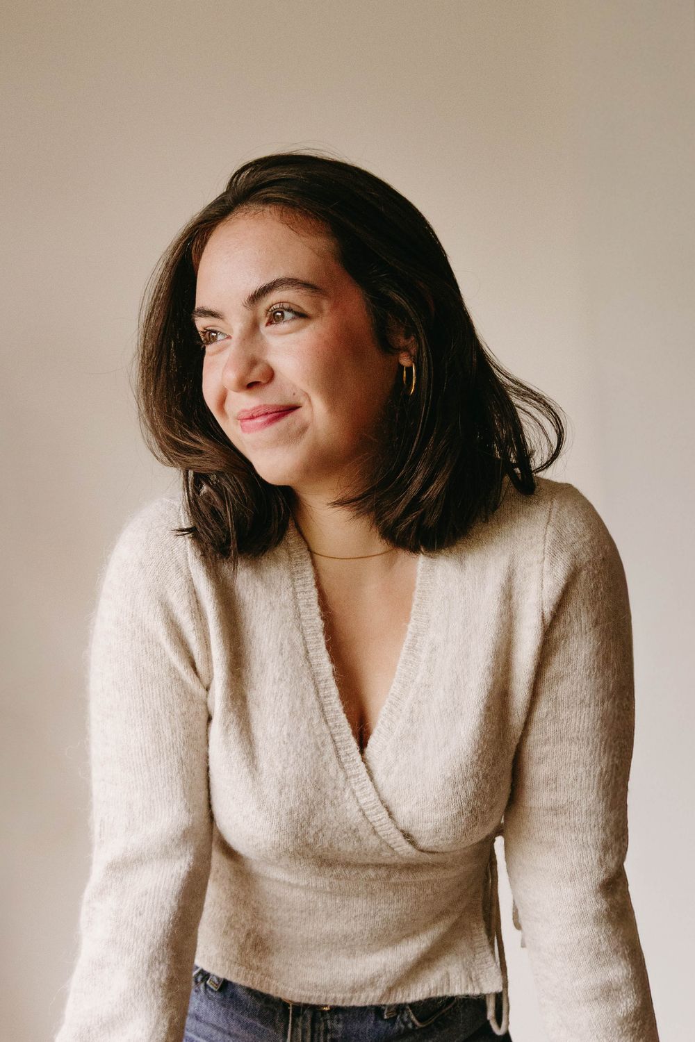 Smiling woman with shoulder-length dark hair wears a cream wrap sweater and jeans, looking off to the side against a neutral backdrop.