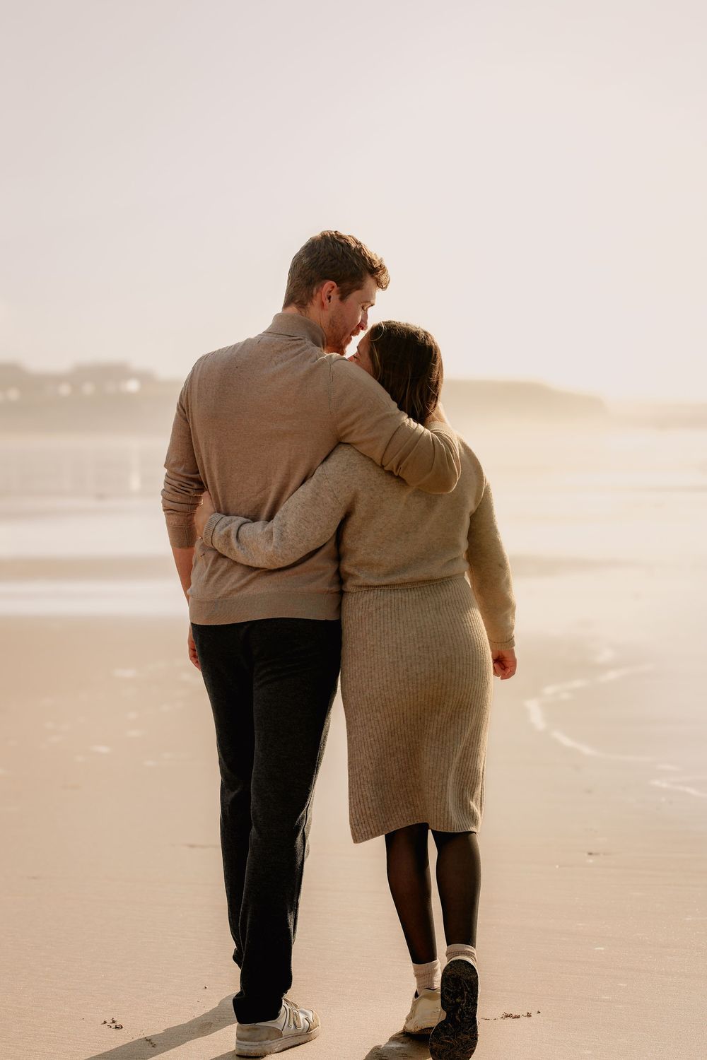 Couple in cozy knitwear walking arm in arm along a sandy beach at golden hour, seen from behind with soft misty ocean light.