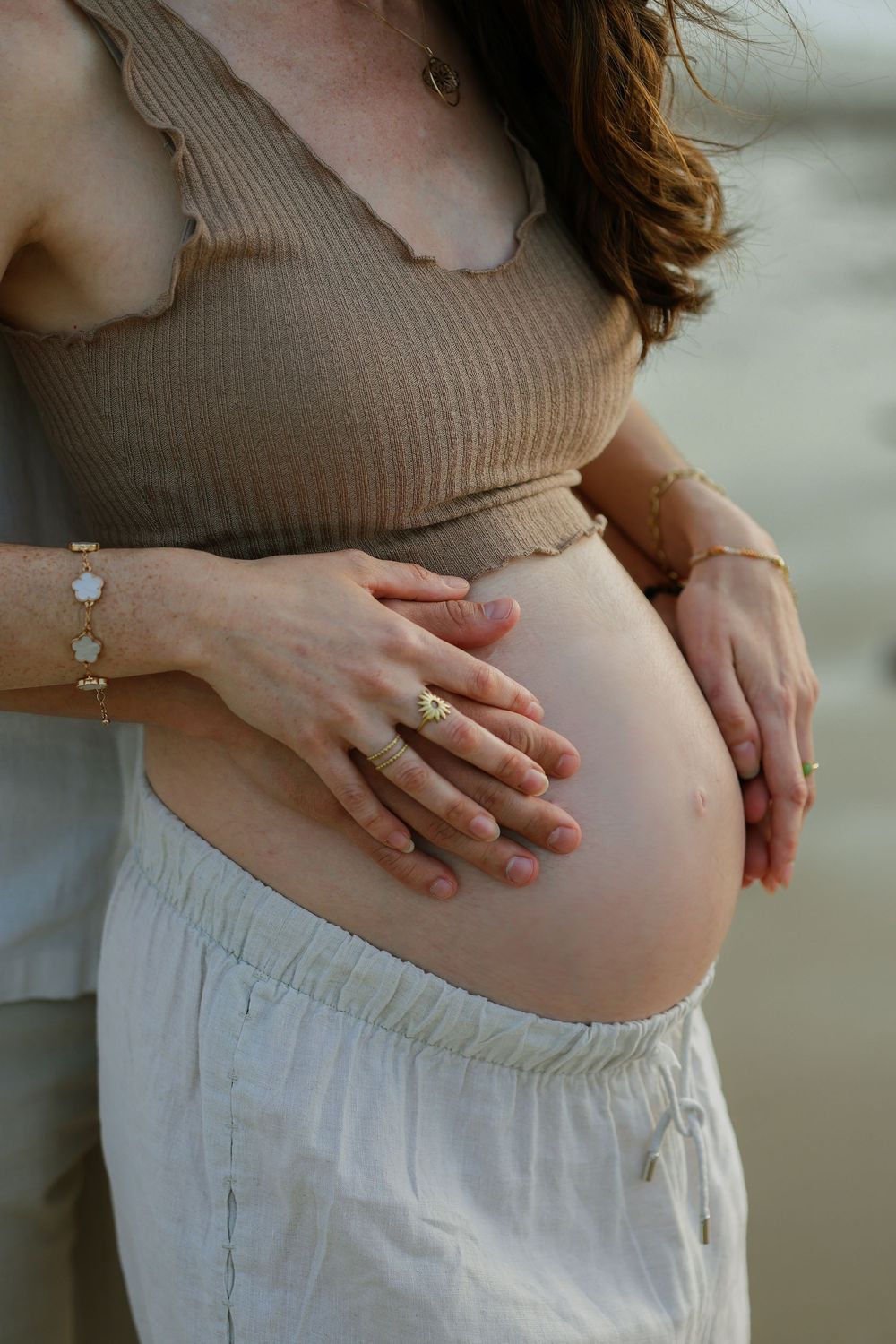Close-up of a pregnant woman in a tan ribbed top and white linen pants cradling her bare baby bump with both hands on a beach.