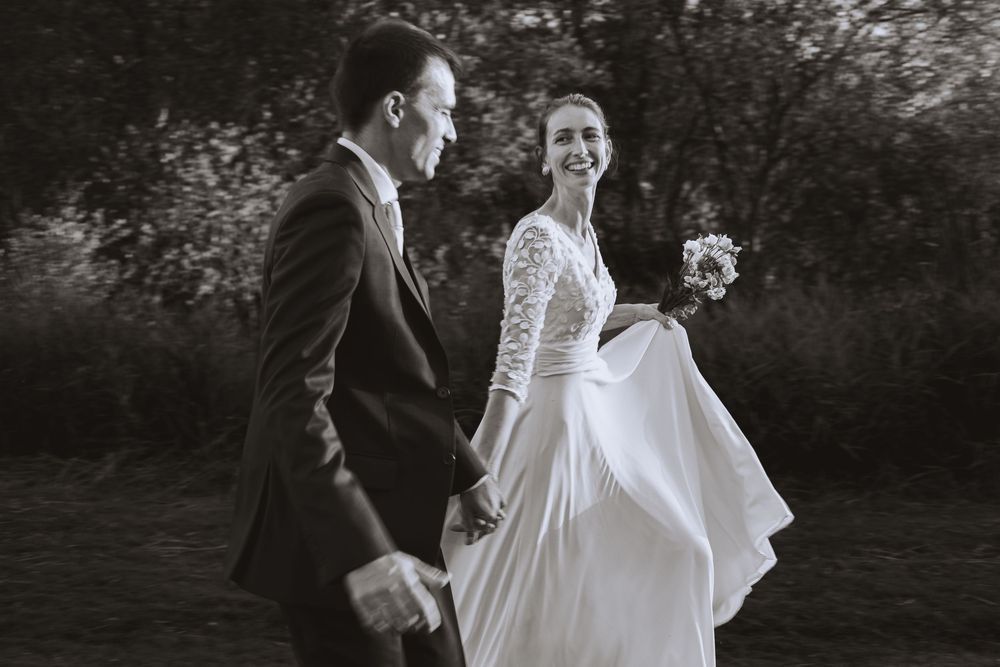 Black and white photo of a smiling bride in a lace-sleeved gown holding a bouquet and walking hand in hand with the groom outdoors.