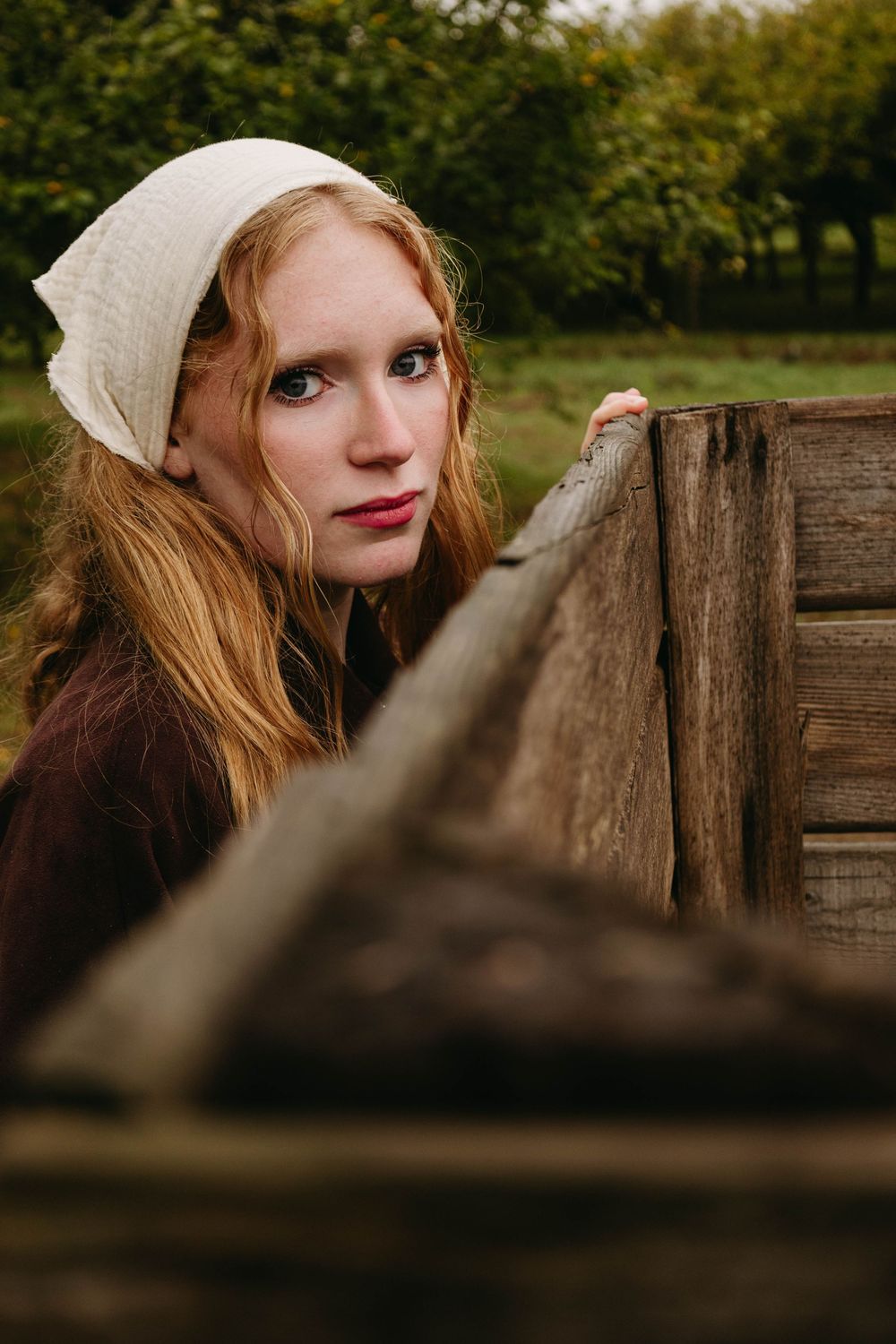 Young woman with long red hair and a white linen headscarf leans on a rustic wooden fence in a green orchard, gazing at the camera.