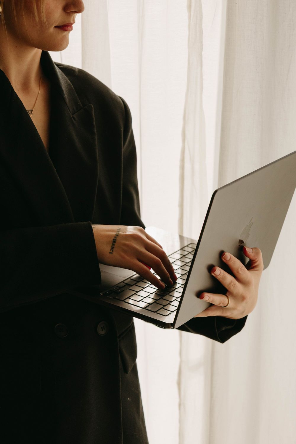 Woman in a black blazer stands by sheer white curtains, typing on a silver laptop held in one hand.